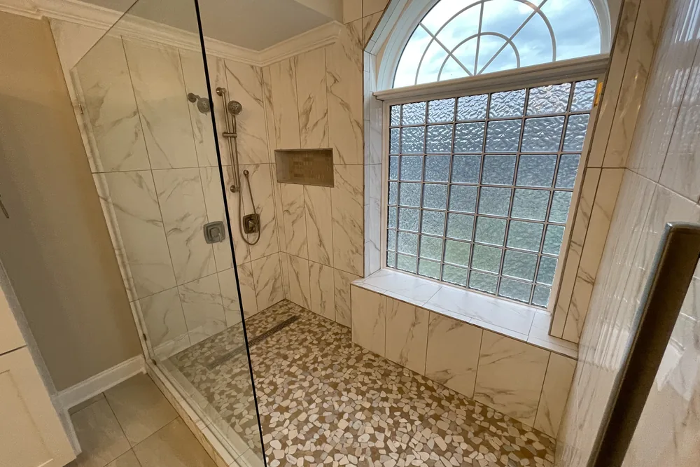Remodeled walk-in shower with marble-look tile, a pebble-tile floor, and a glass-block window.