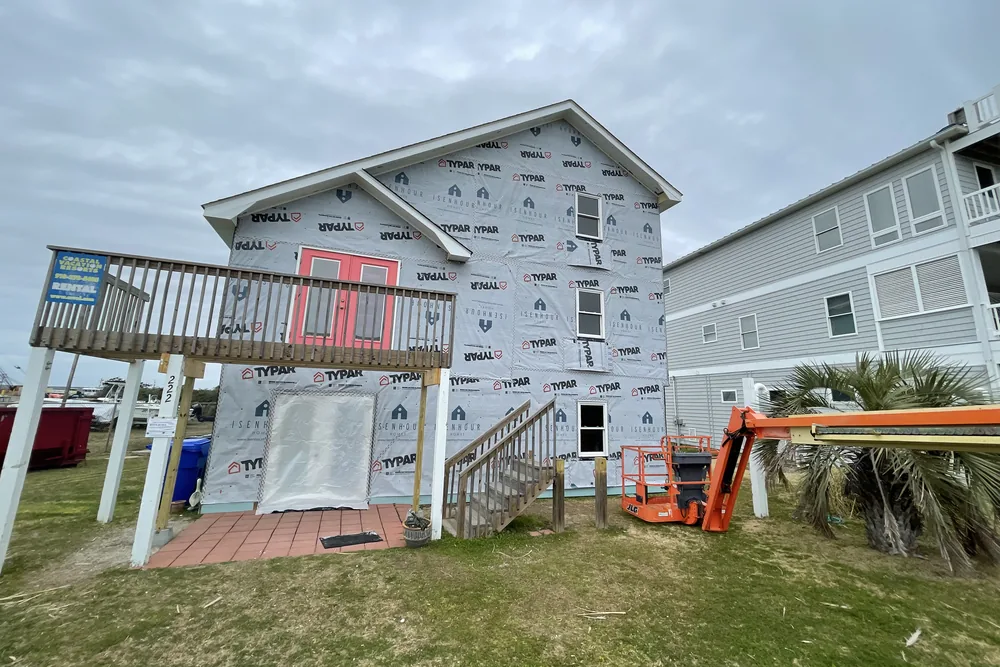 In progress: rear of a raised beach house in Caswell Beach, fully wrapped in house wrap, with a boom lift beside it.