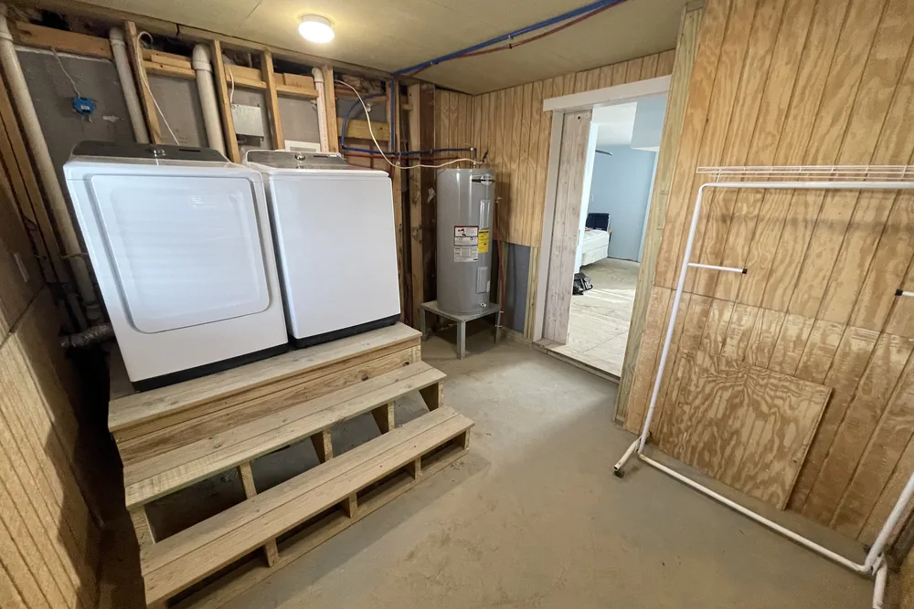 Before: washer and dryer on a wood platform beside open wall framing and a water heater, in Oak Island.